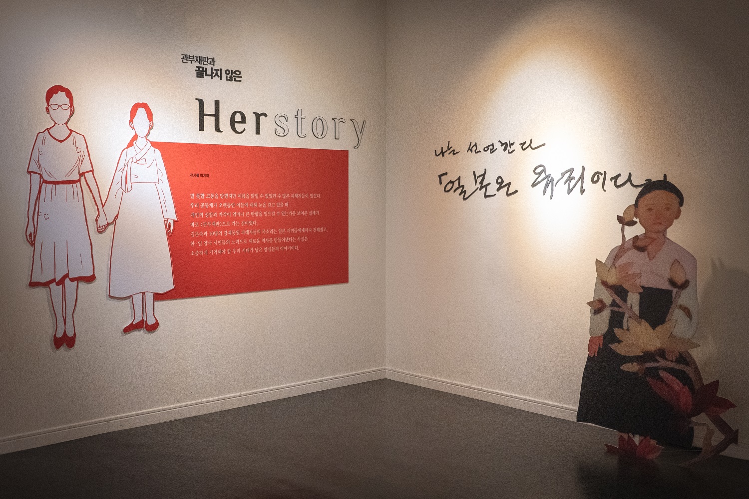&lsquo;관부재판과 끝나지 않은 Herstory&rsquo; 전시를 마치며 쓴 글과 손을 맞잡은 두 여성 그림이 있는 벽이다. 오른쪽 벽에는 &ldquo;나는 선언한다 「일본은 유죄이다」&rdquo;라는 손글씨 문구와 꽃을 든 소녀 그림 입간판이 놓여 있다.