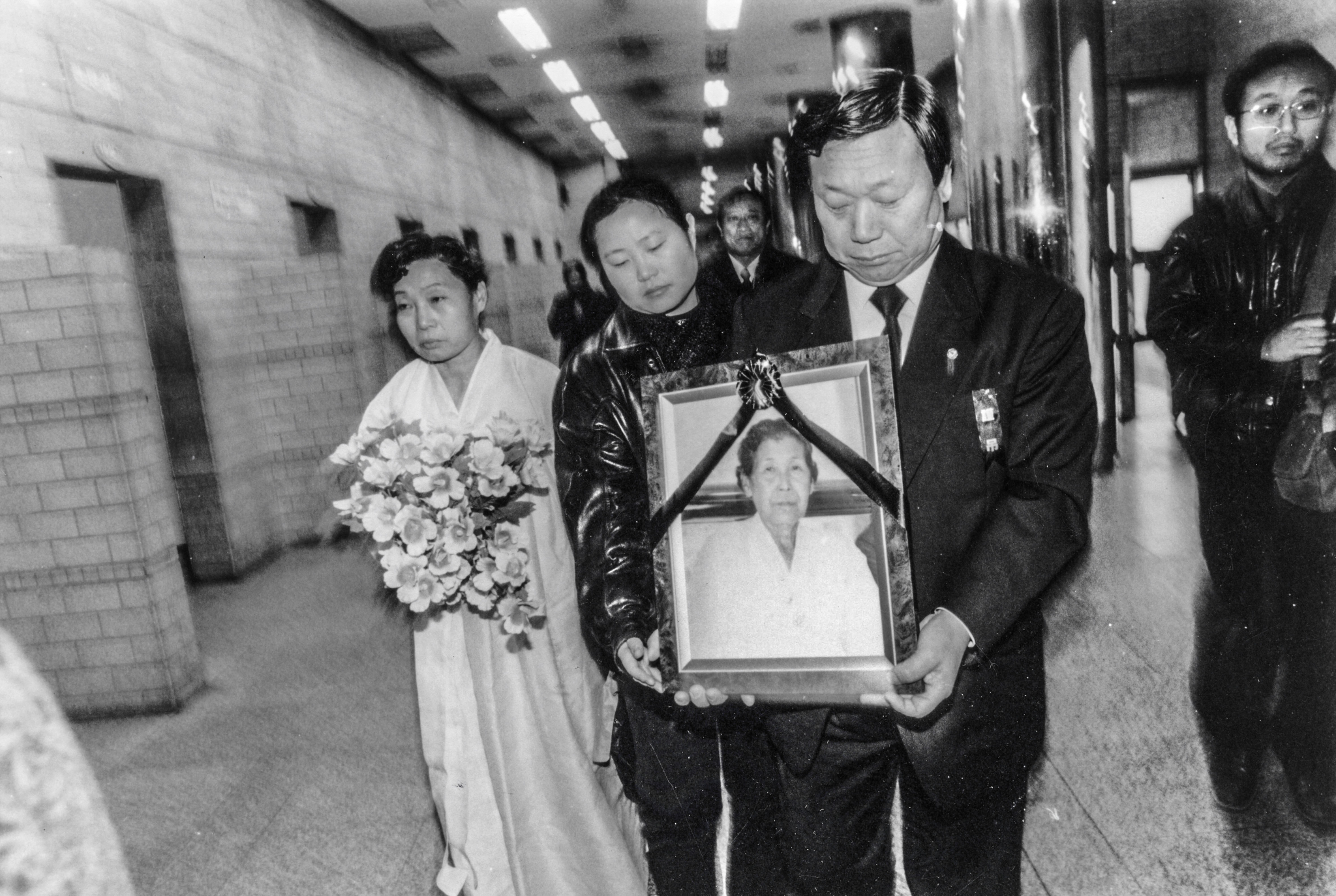 김학순 할머니 장례식 흑백 사진이다. 검은 띠를 두른 김학순 할머니의 영정을 정장 차림의 참석자가 두 손으로 들고 복도를 걸어가고, 옆에서 다른 참석자가 영정을 함께 받치고 있다. 그 왼쪽에는 흰 한복 차림의 참석자가 꽃다발을 안고 함께 걷고 있으며, 뒤로 여러 사람이 따르고 있다.