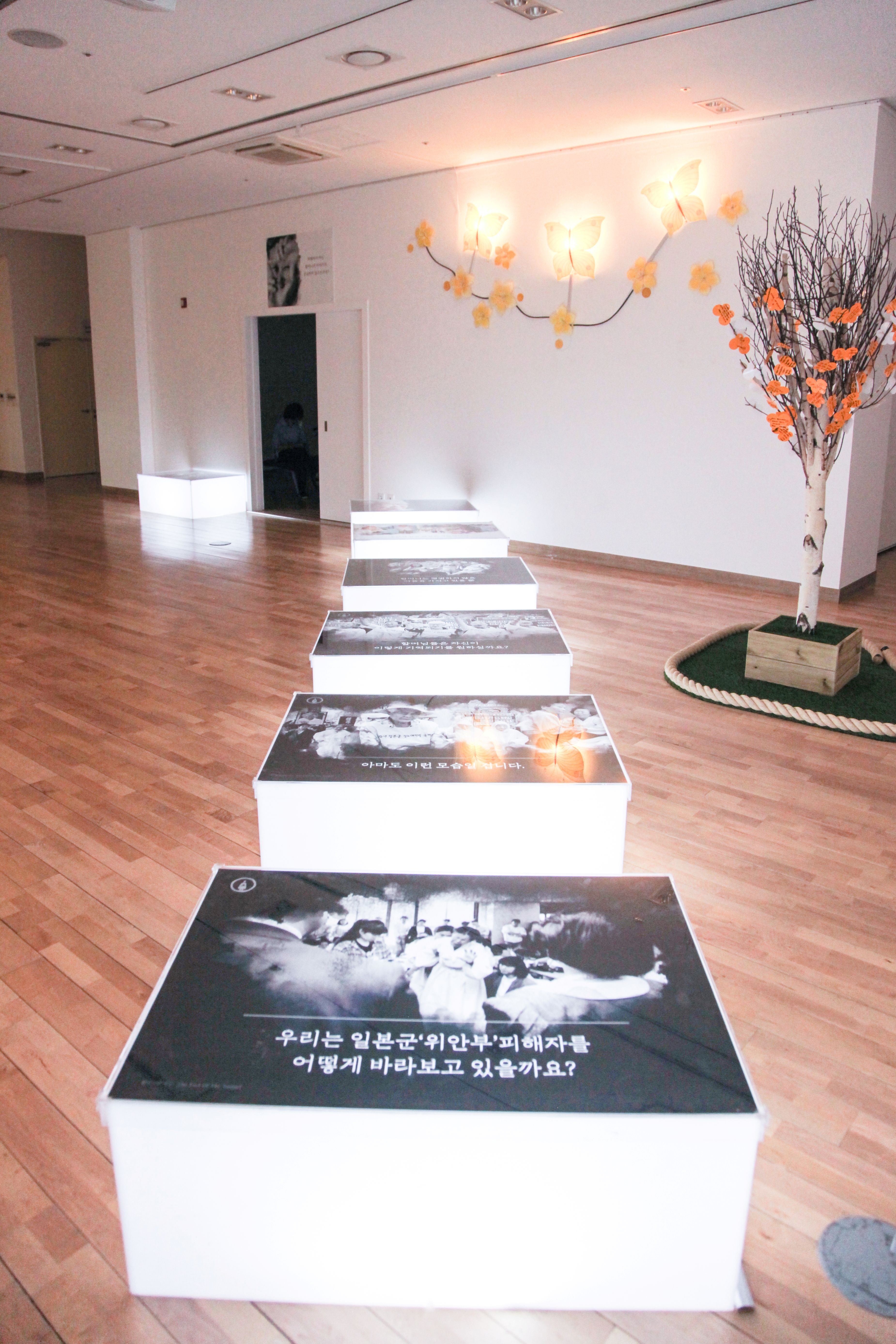 바닥에 일렬로 놓인 조명 상자 위에 사진 패널이 이어져 있고, 맨 앞 패널에 &ldquo;우리는 일본군&lsquo;위안부&rsquo;피해자를 어떻게 바라보고 있을까요?&rdquo;라는 질문이 적혀 있다. 안쪽 벽에는 나비 모양 조명이, 오른쪽에는 메시지 카드가 매달린 나무가 있다.