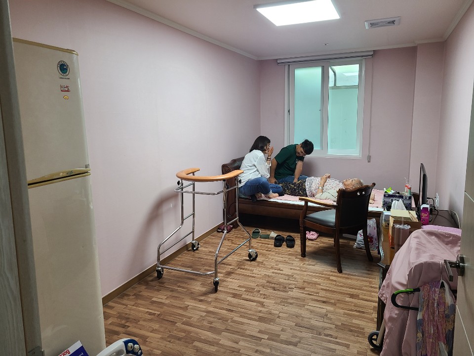 방 입구에서 바라본 모습이다. 왼쪽에 냉장고와 보행 보조기가 있고, 안쪽 창가 침대에 누운 할머니 곁에 두 사람이 앉아 있다.