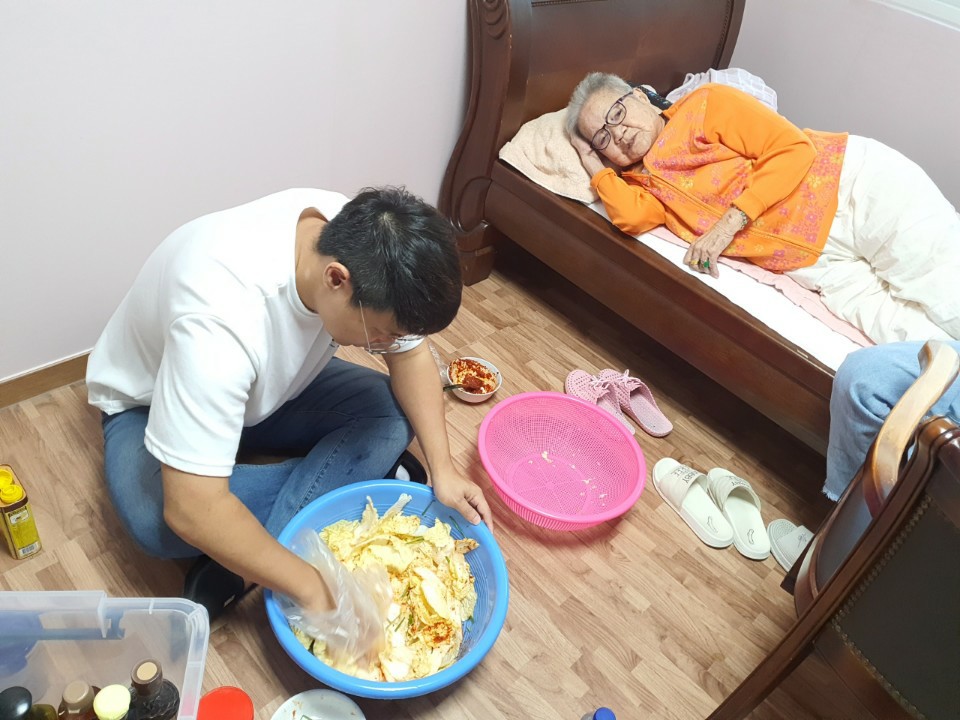 속리산 이옥선 할머니 방바닥에서 파란 대야에 배추 겉절이를 버무리고 있고, 침대에 누운 할머니가 이를 지켜보고 있다.
