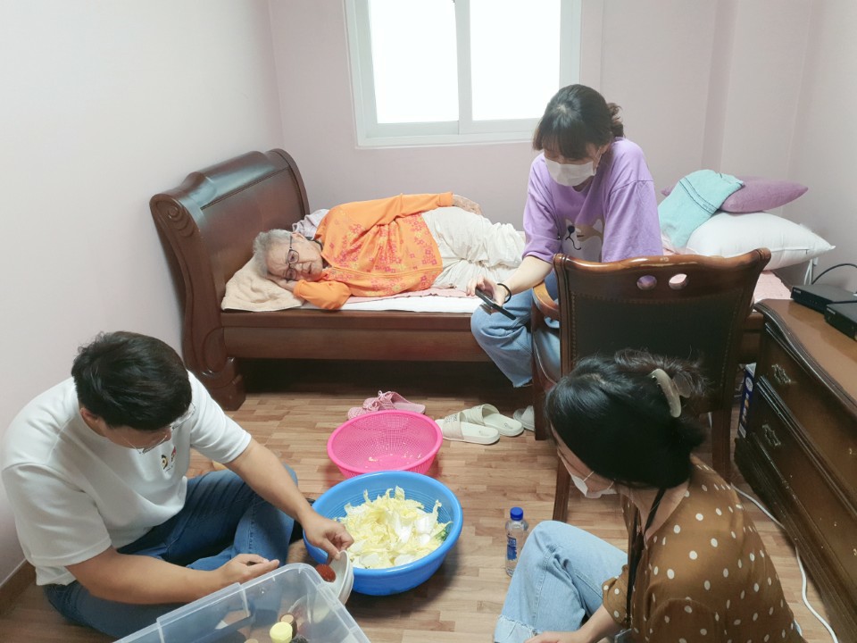 침대에 누운 속리산 이옥선 할머니 곁에서 세 사람이 바닥과 의자에 둘러앉아 함께 겉절이를 만들고 있다.