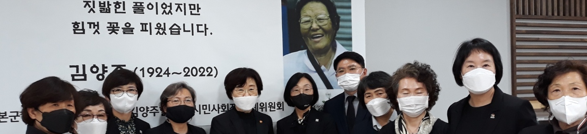 김양주 할머니 장례식장에서 검은 옷을 입은 조문객들이 나란히 서서 찍은 사진이다. 뒤편 현수막에 웃고 있는 김양주 할머니의 사진과 함께 &ldquo;짓밟힌 풀이었지만 힘껏 꽃을 피웠습니다. 김양주(1924~2022)&rdquo;라는 문구가 적혀 있다.