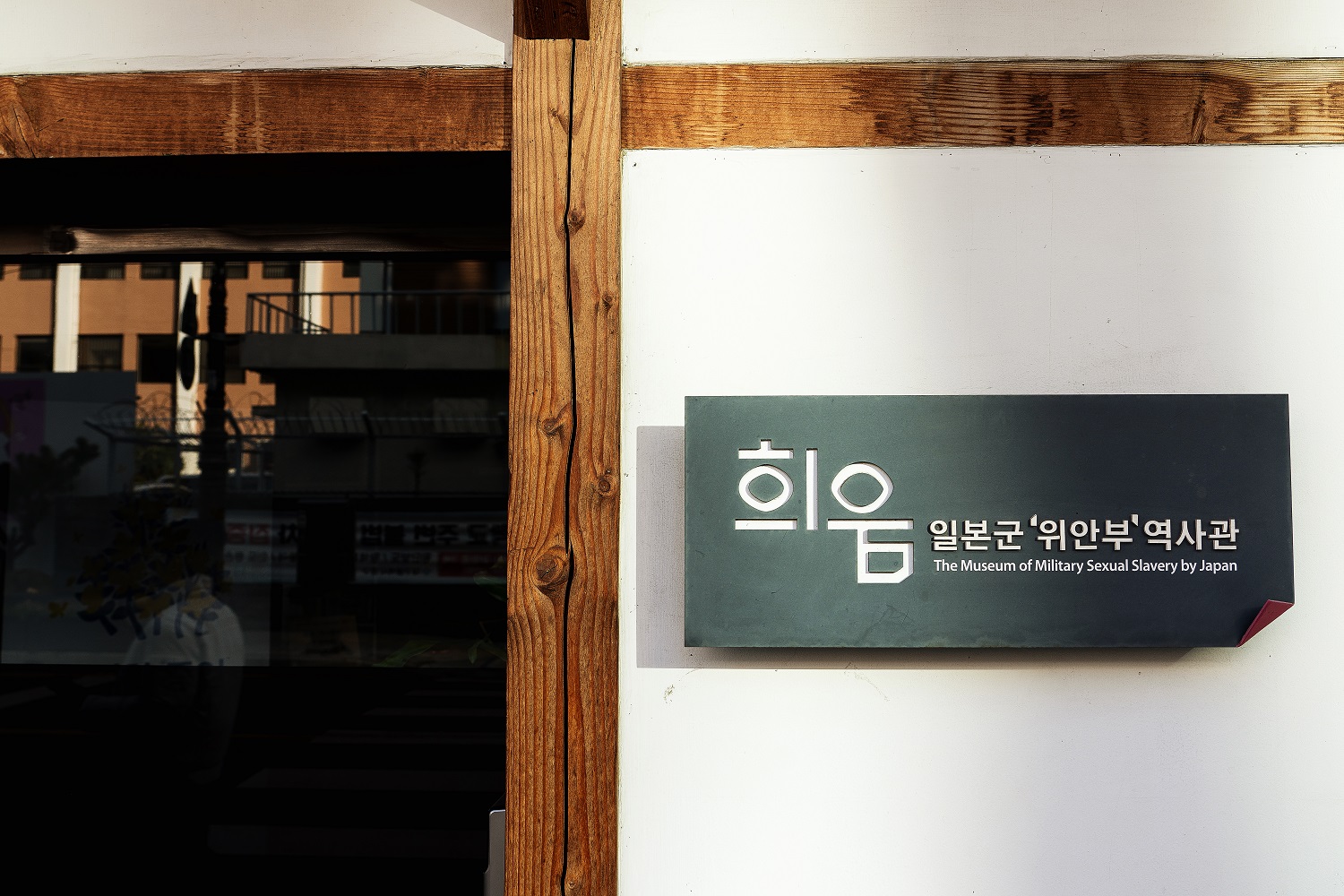 희움 일본군&lsquo;위안부&rsquo;역사관의 간판이다. 나무 기둥이 있는 흰 벽에 국문 이름과 영문 이름(The Museum of Military Sexual Slavery by Japan)이 새겨진 짙은 회색 간판이 걸려 있다.