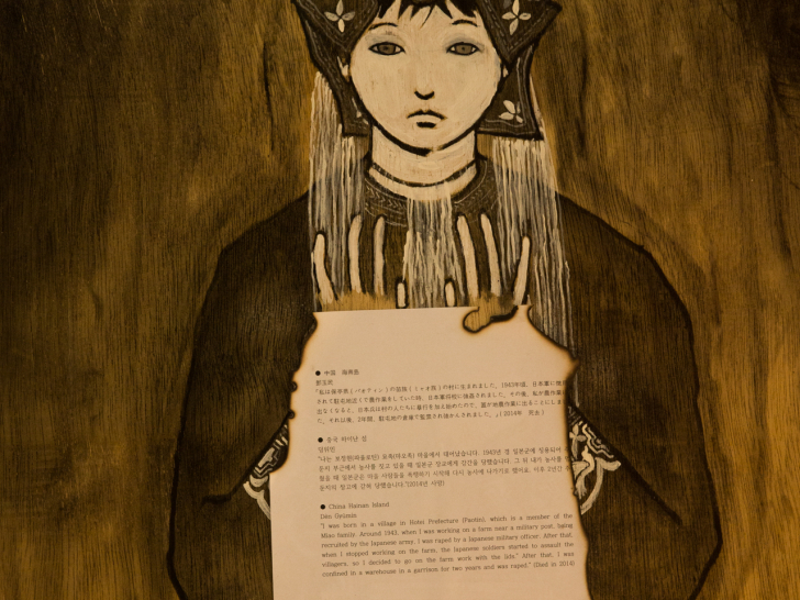 &lsquo;일본군 성노예 연작&rsquo; 중 한 인물을 가까이에서 찍은 모습이다. 전통 머리 장식을 한 여성이 가장자리가 불에 그을린 증언 종이를 들고 있으며, 종이에는 중국 하이난섬 피해 여성의 증언이 일본어, 한국어, 영어로 적혀 있다.