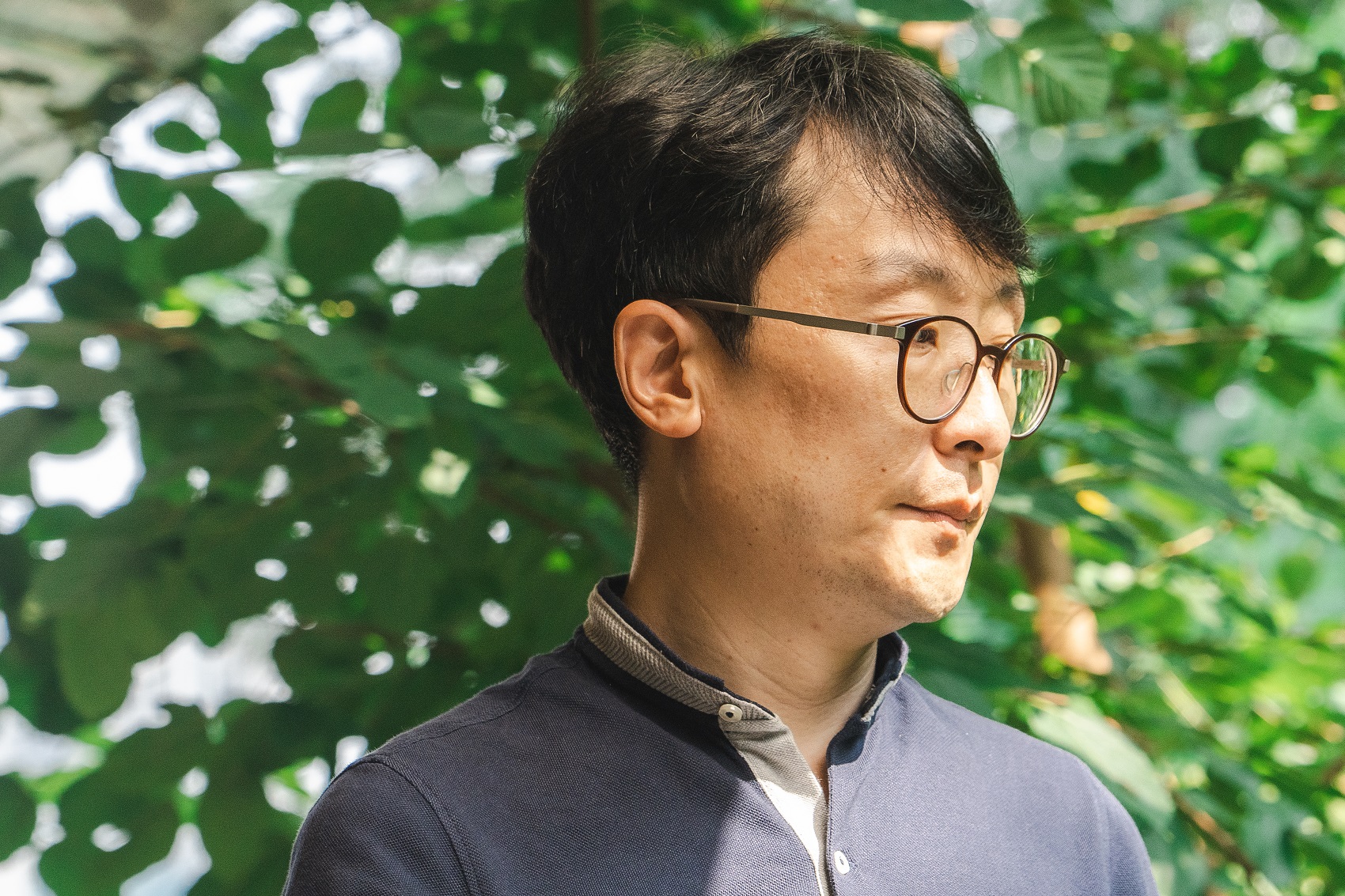 푸른 나뭇잎을 배경으로 안경을 쓴 박문칠 감독이 옆을 바라보고 있는 인물 사진