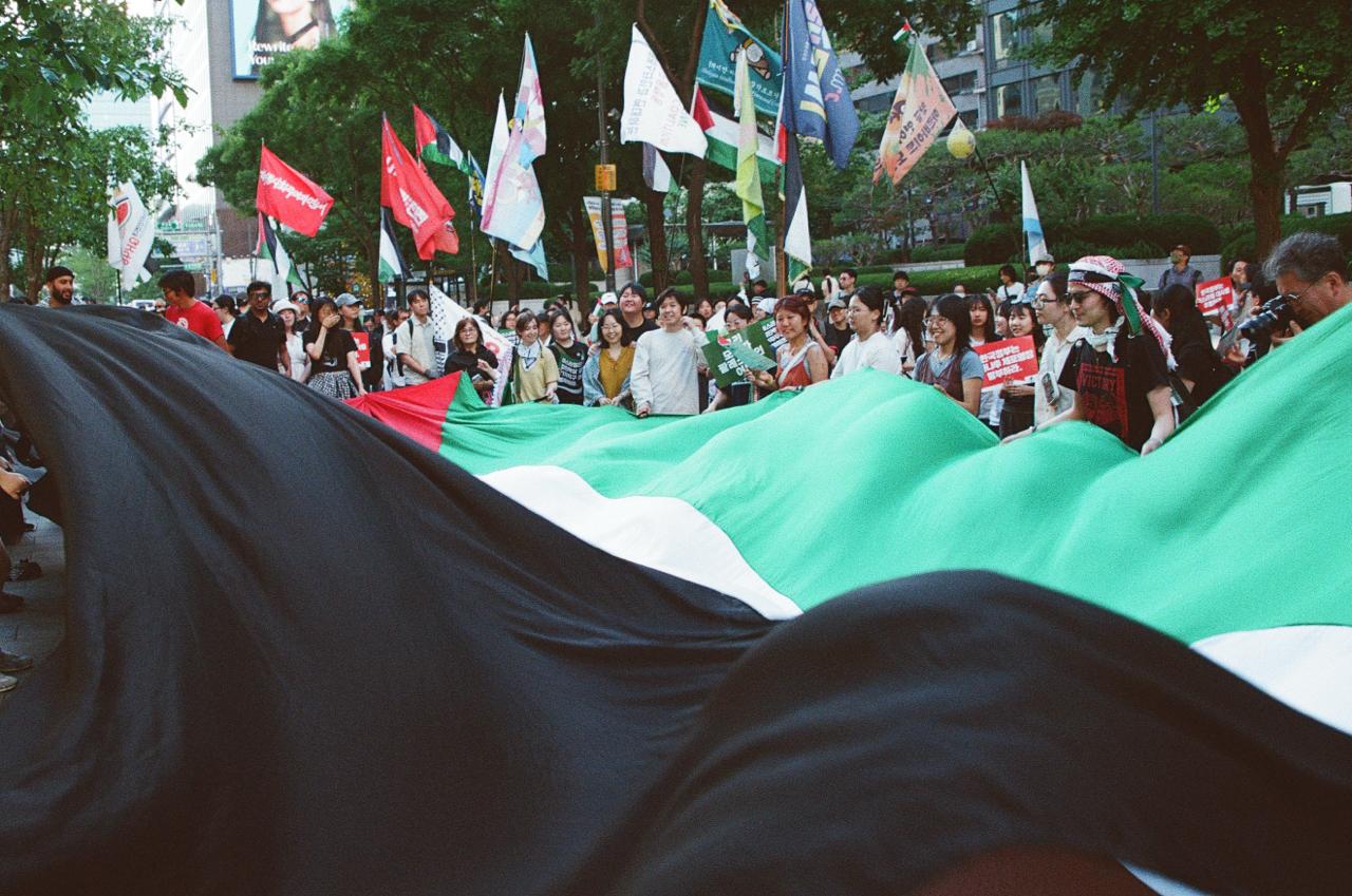 (사진 4) 2차 나포 후 항해에서 돌아온 직후 김동현, 이승준, 해초 항해자들이 팔레스타인 연대 집회에서 시민들이 접은 종이 돛단배를 팔레스타인 국기를 따라 흐르게 하는 퍼포먼스를 하고 있다. (사진 제공: 해초)